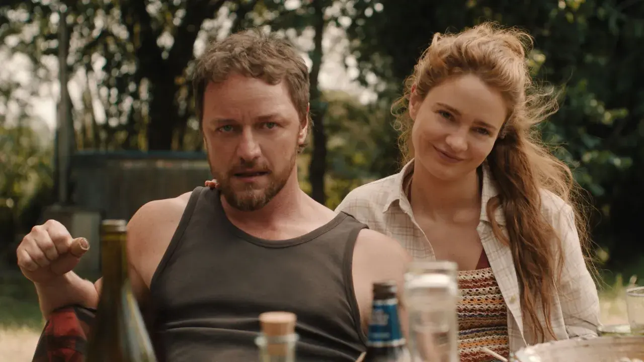 Una coppia sorride, un uomo con barba e canottiera, una donna con capelli rossi. La scena evoca un'atmosfera da "speak no evil recensione".