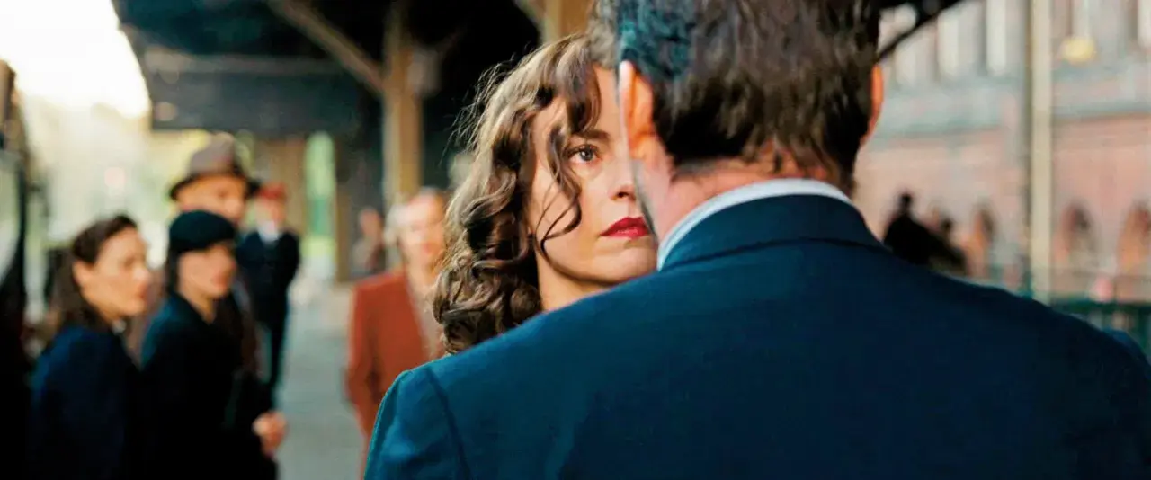 Una donna con labbra rosse e capelli ricci guarda intensamente un uomo, il segreto del suo volto trama. Sullo sfondo, persone sfocate in una stazione.