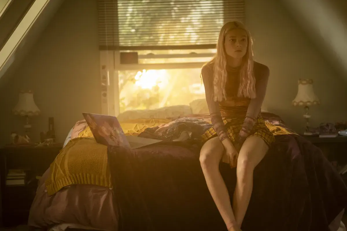 Una ragazza bionda con un vestito a quadri siede sul letto, illuminata dalla luce del tramonto. Una scena che fa pensare a una recensione di Euphoria.