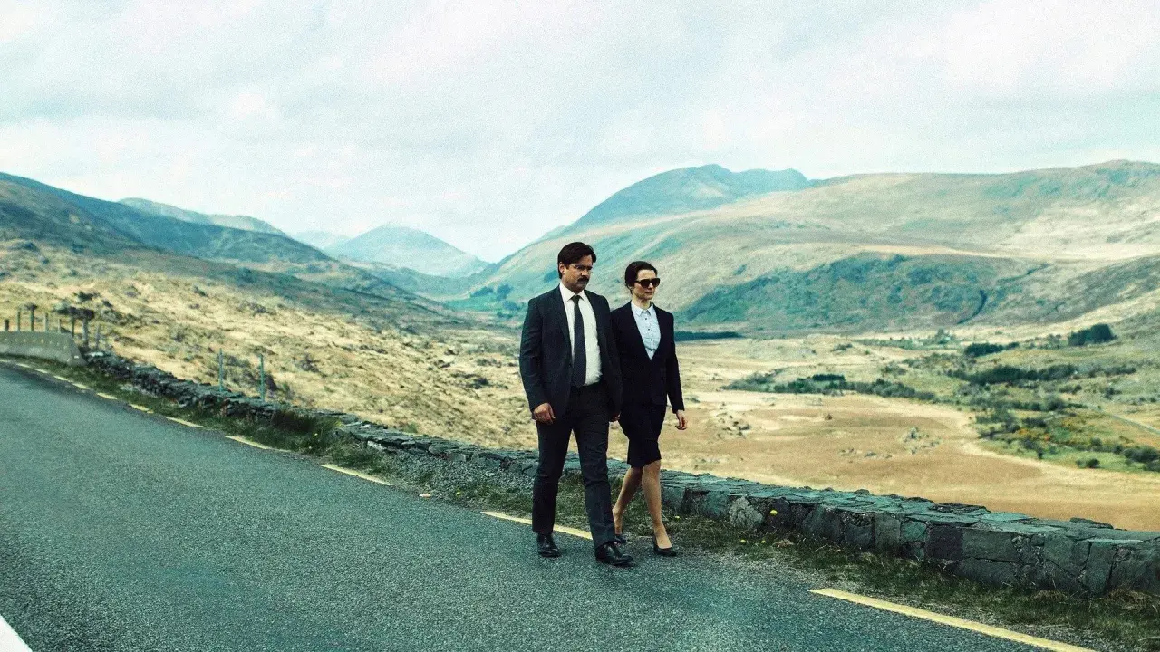 Coppia in abiti eleganti cammina su strada panoramica, evocando il significato di "The Lobster" e la ricerca di un legame in un paesaggio desolato.