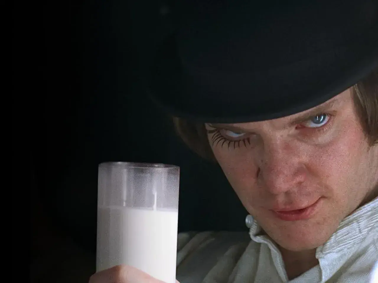 Un uomo con un cappello nero e ciglia finte tiene un bicchiere di latte, rompendo la quarta parete con uno sguardo intenso.