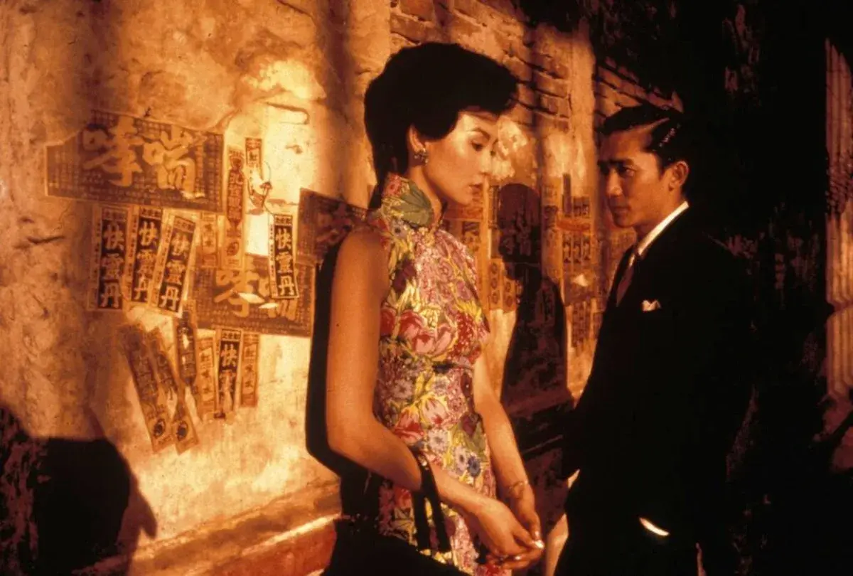 In the mood for love spiegazione: una donna in cheongsam fiorito e un uomo in abito scuro si guardano con intensità, sullo sfondo di un muro con scritte cinesi.