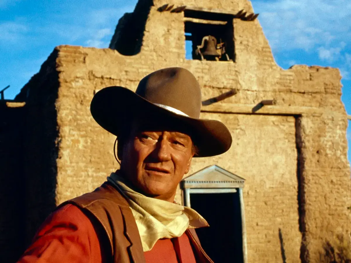 John Wayne, icona del West, con cappello e bandana, davanti a una chiesa diroccata. Ricorda le sue frasi western famose.