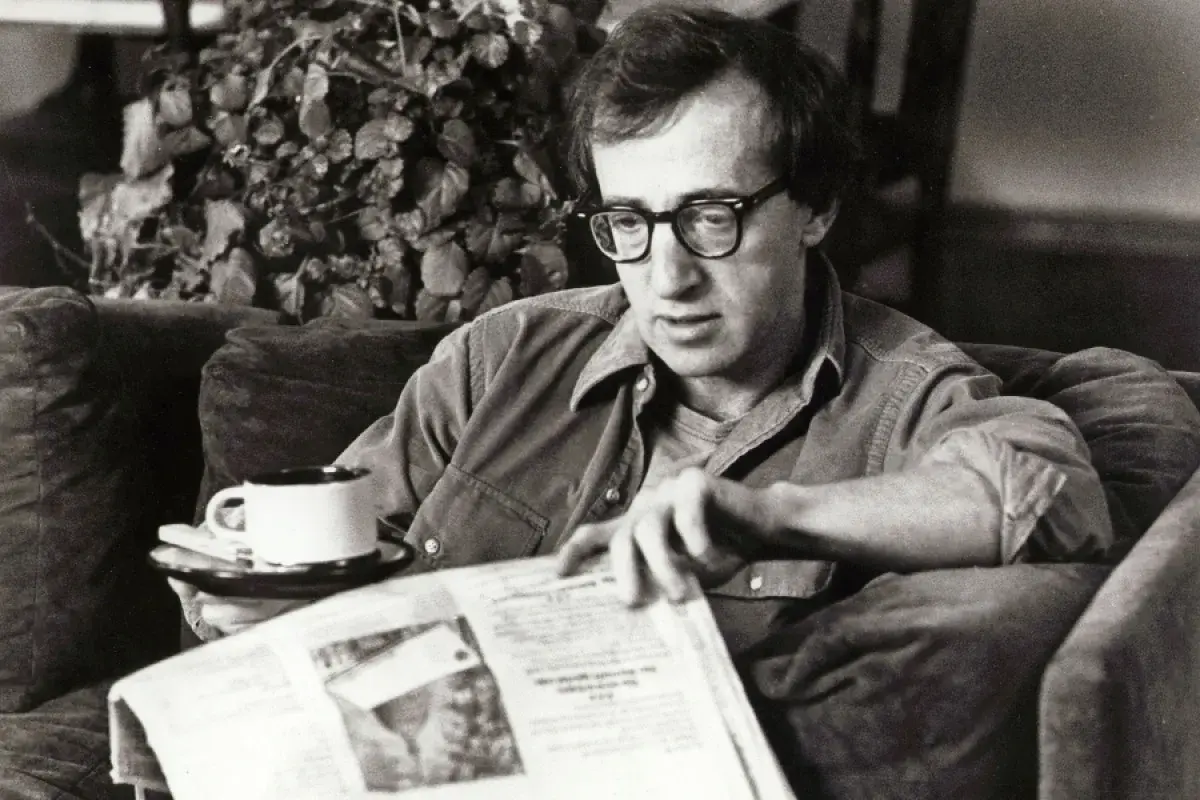 Woody Allen, con occhiali e camicia, legge un giornale con una tazza di caffè, forse pensando al suo prossimo Oscar.