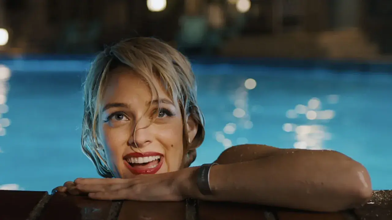 Una donna sorride dall'acqua di una piscina, con luci sfocate sullo sfondo. Una scena che ricorda "Under the Silver Lake".
