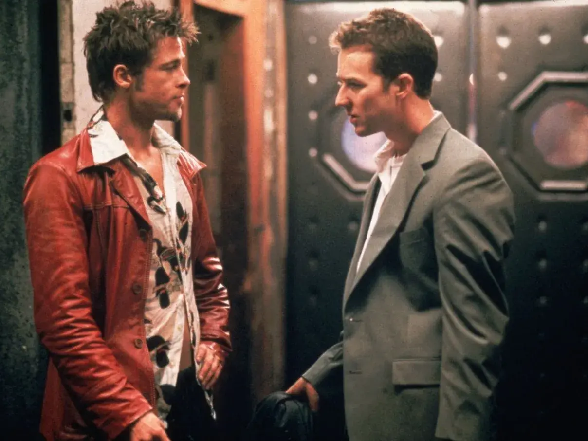 Il narratore di Fight Club, con la sua giacca rossa, parla con un uomo in completo grigio.