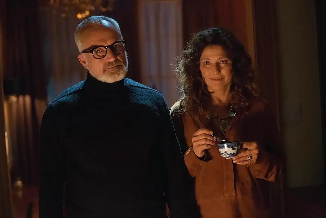 Coppia anziana in un interno buio, lui con occhiali e dolcevita nero, lei sorride con una tazza in mano. Sembra il **get out finale**.