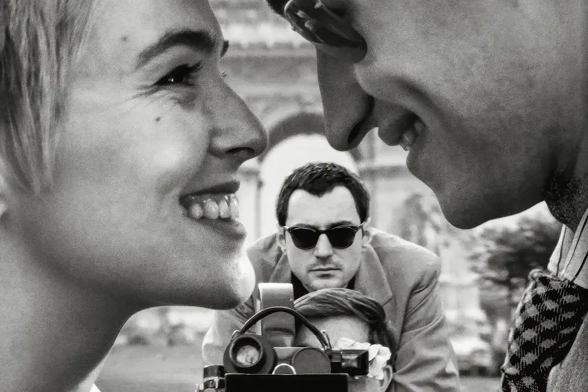 Primo piano di una donna sorridente, con un uomo che la guarda e un altro uomo con occhiali da sole che filma. Un omaggio ai registi importanti.