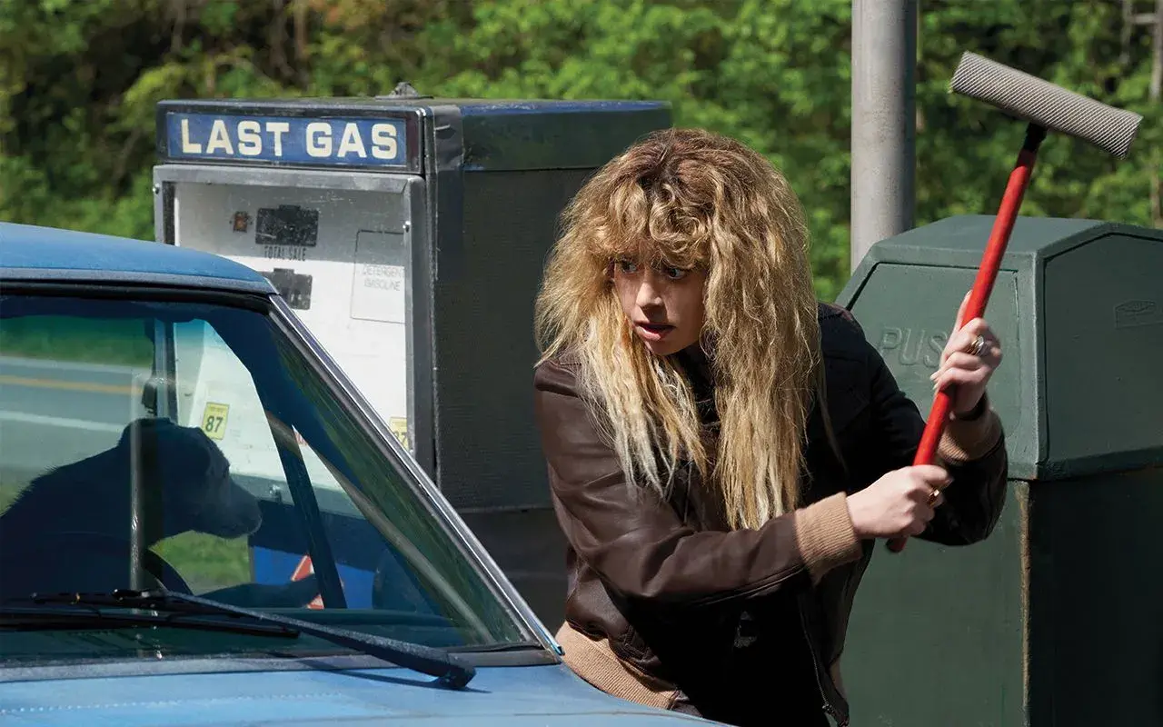 La protagonista di Poker Face, Nadia Vulvokov, con i suoi capelli biondi spettinati, impugna un tergivetro rosso davanti a una pompa di benzina "Last Gas".