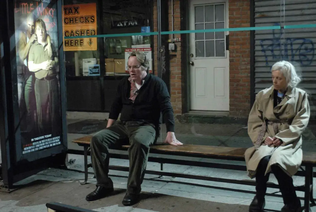 Uomo e donna seduti su una panchina, vicino a un poster di "Little Winky". Una scena che evoca la complessità di "Synecdoche, New York".