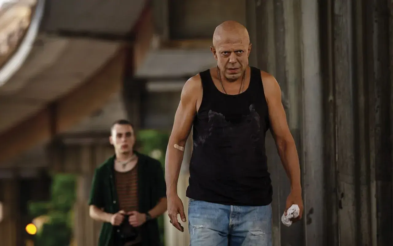 Due uomini, uno calvo e con un cerotto sul braccio, camminano in un ambiente urbano. Scena dal film "Adagio".