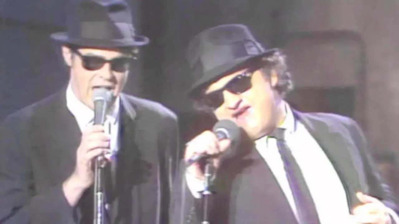 Dan Aykroyd giovane e John Belushi sul palco, entrambi con cappello e occhiali da sole, cantano al microfono.