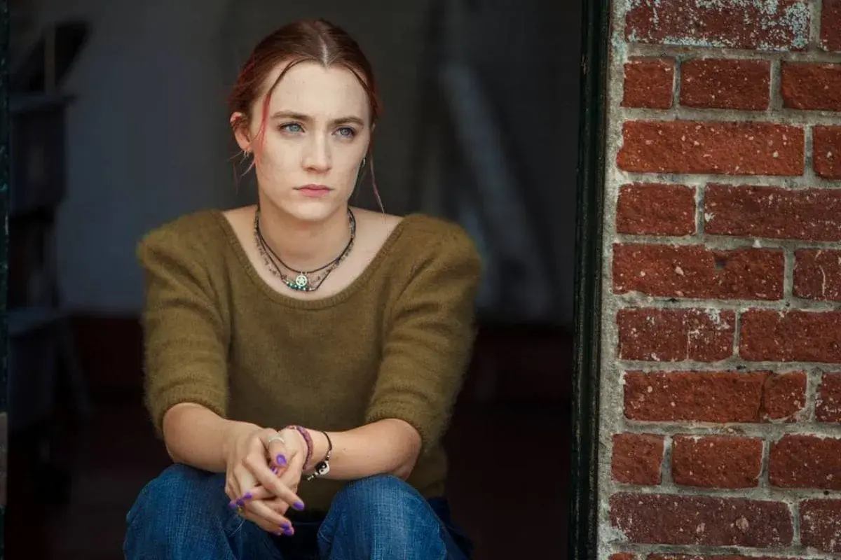 Ragazza pensierosa, con maglione verde e jeans, seduta vicino a un muro di mattoni rossi. Una scena che ricorda la recensione di Lady Bird.