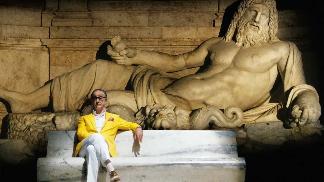Un uomo in giacca gialla seduto su una panchina di marmo, con una statua antica alle spalle. Un'immagine che evoca la grande bellezza, spiegazione visiva dell'arte e del tempo.