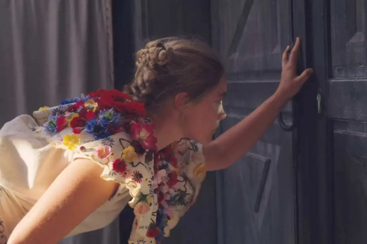 Una ragazza con un vestito fiorito, capelli raccolti, spinge una porta scura. Un'immagine che evoca la recensione di Midsommar.