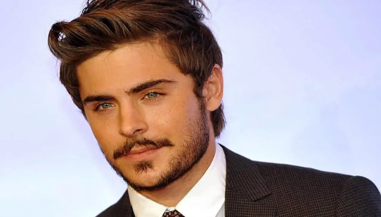 Primo piano di Zac Efron giovane, con sguardo intenso e barba curata, indossa una giacca scura.