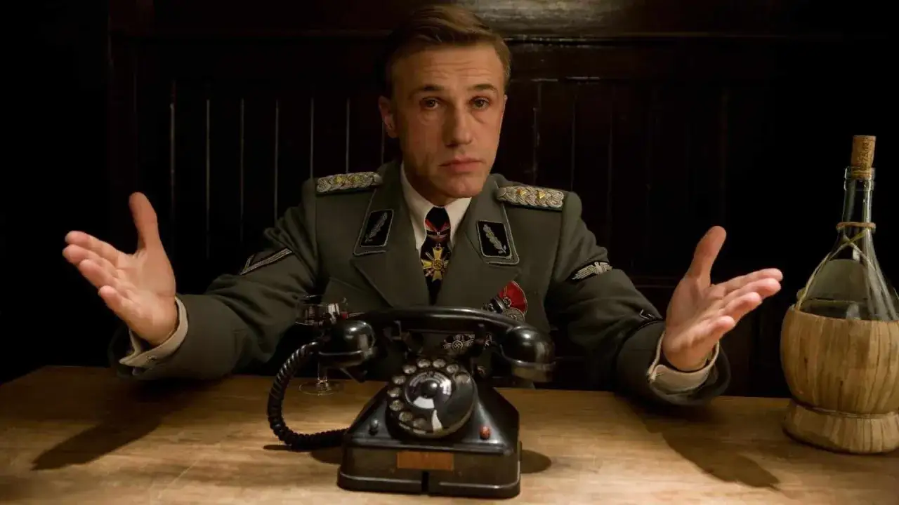 Ufficiale nazista con le mani aperte, un telefono d'epoca e una bottiglia di vino. Il suo sguardo è intenso, quasi a voler spiegare il significato di
