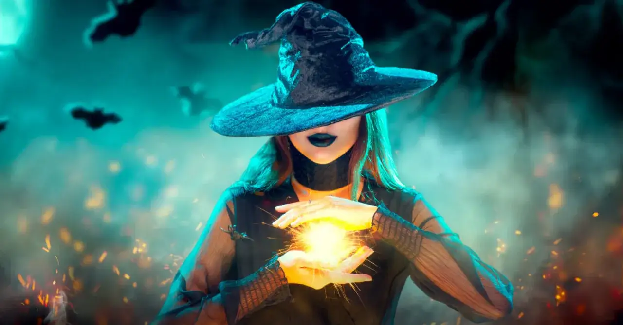 La strega hazel, con cappello nero e labbra scure, evoca una sfera di luce tra le mani. Pipistrelli volano nel cielo nebbioso.
