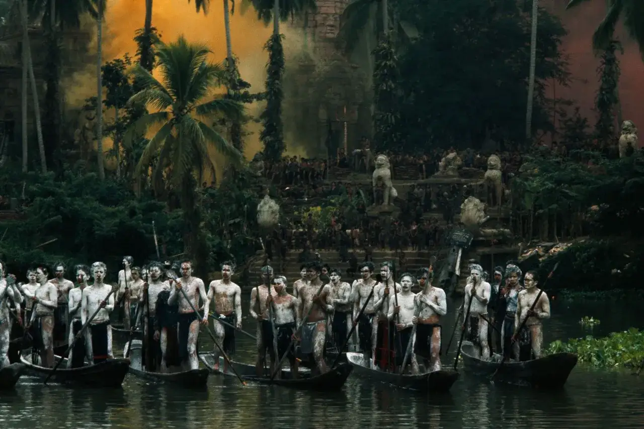 Uomini in canoe, con corpi dipinti, remano verso una scena caotica con fumo e folla, evocando il significato di 