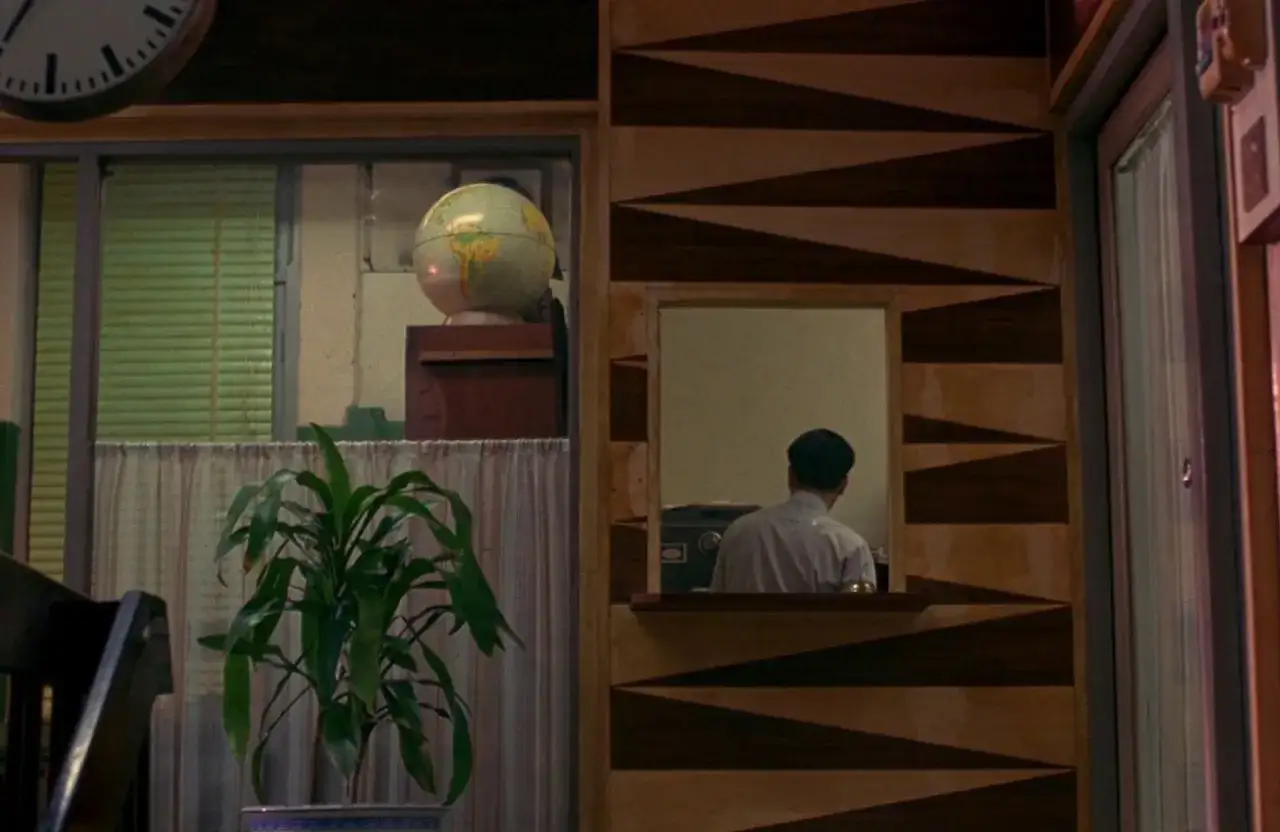 Un uomo dietro un bancone, un globo terrestre e una pianta. Atmosfera malinconica, perfetta per le recensioni di 