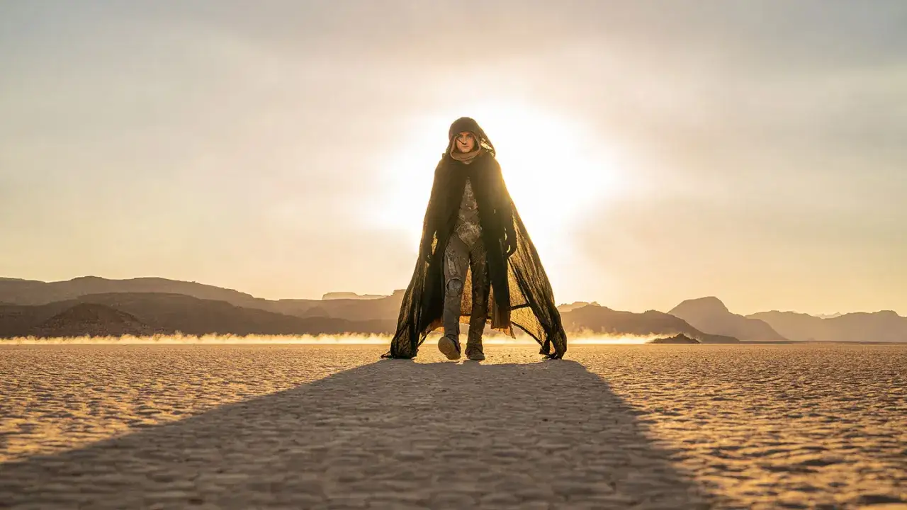 Un personaggio con mantello scuro cammina in un deserto arido, sotto un sole accecante. Le dune recensioni sembrano infinite.