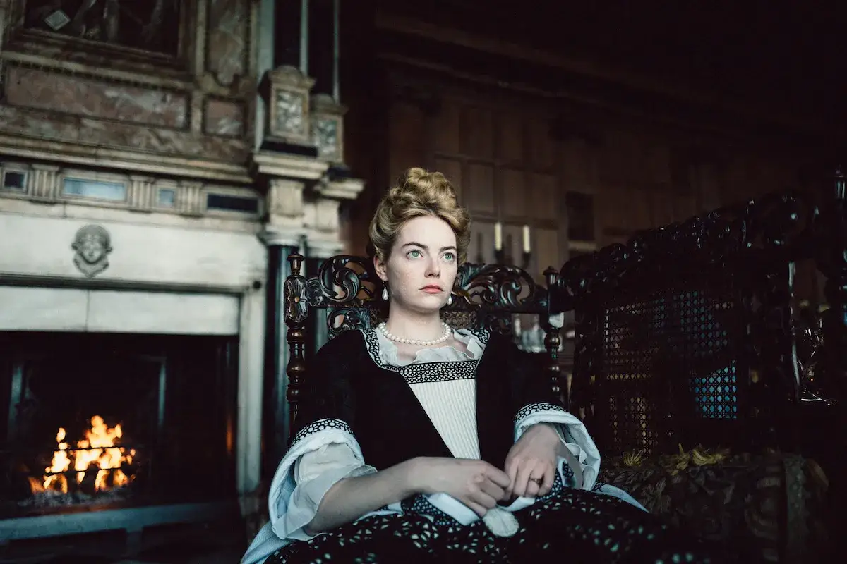 La favorita recensione: Emma Stone, elegante in abito d'epoca, siede su una sedia intagliata, con un camino acceso sullo sfondo.