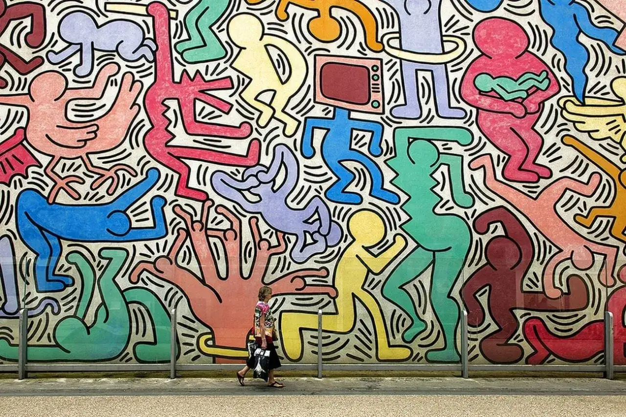 Un mur vibrante di figure stilizzate colorate, un'esplosione di gioia e movimento nello stile inconfondibile di Keith Haring. Una donna cammina ai suoi piedi.