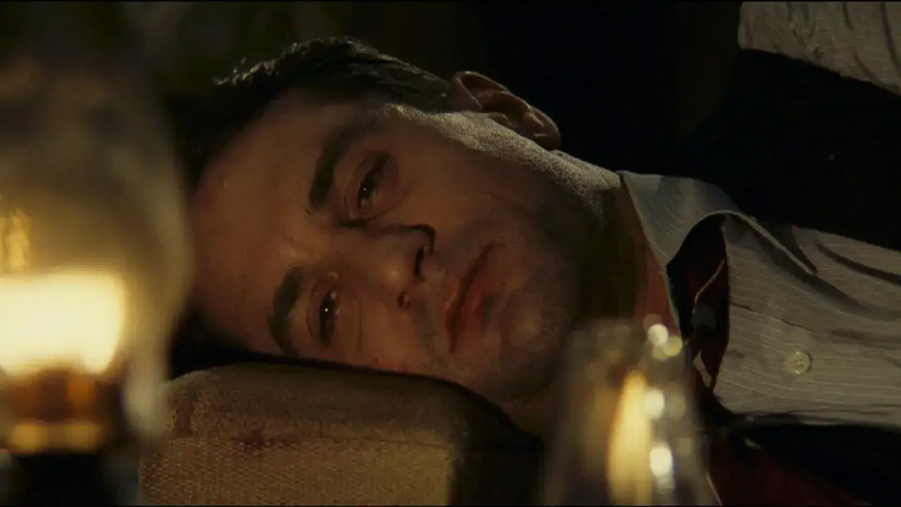 Un uomo giace esausto, con lo sguardo perso nel vuoto. La sua espressione suggerisce una profonda malinconia, quasi una spiegazione finale, come in