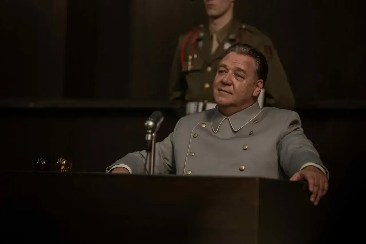 Un uomo in uniforme grigia siede a un podio, con un microfono davanti. Un soldato in uniforme militare è dietro di lui. La scena evoca un'atmosfera da processo, forse legata a un film su Norimberga.
