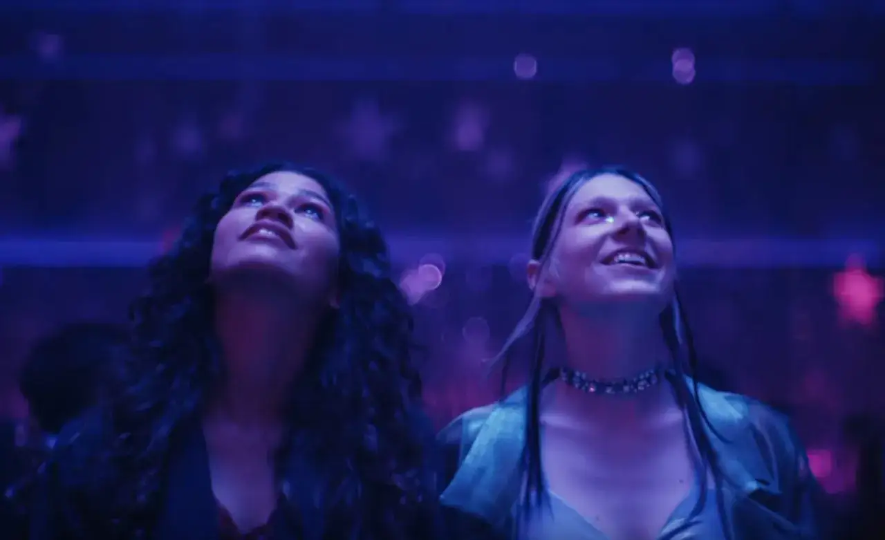 Due ragazze guardano in alto, immerse in luci viola e glitter. Un momento di pura euforia, come in una recensione di Euphoria.