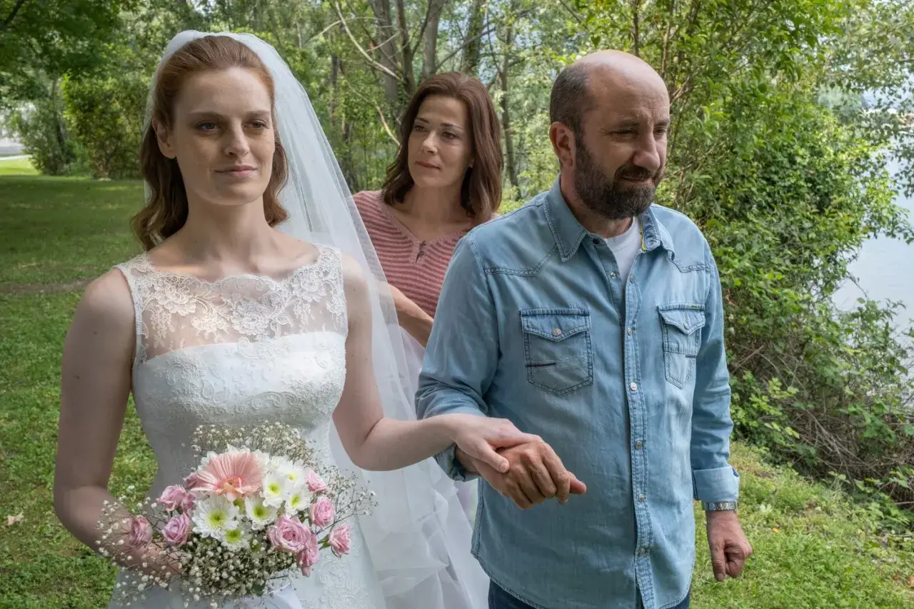 Sposa con velo e bouquet, accompagnata da un uomo e una donna, in un'atmosfera che ricorda il finale di
