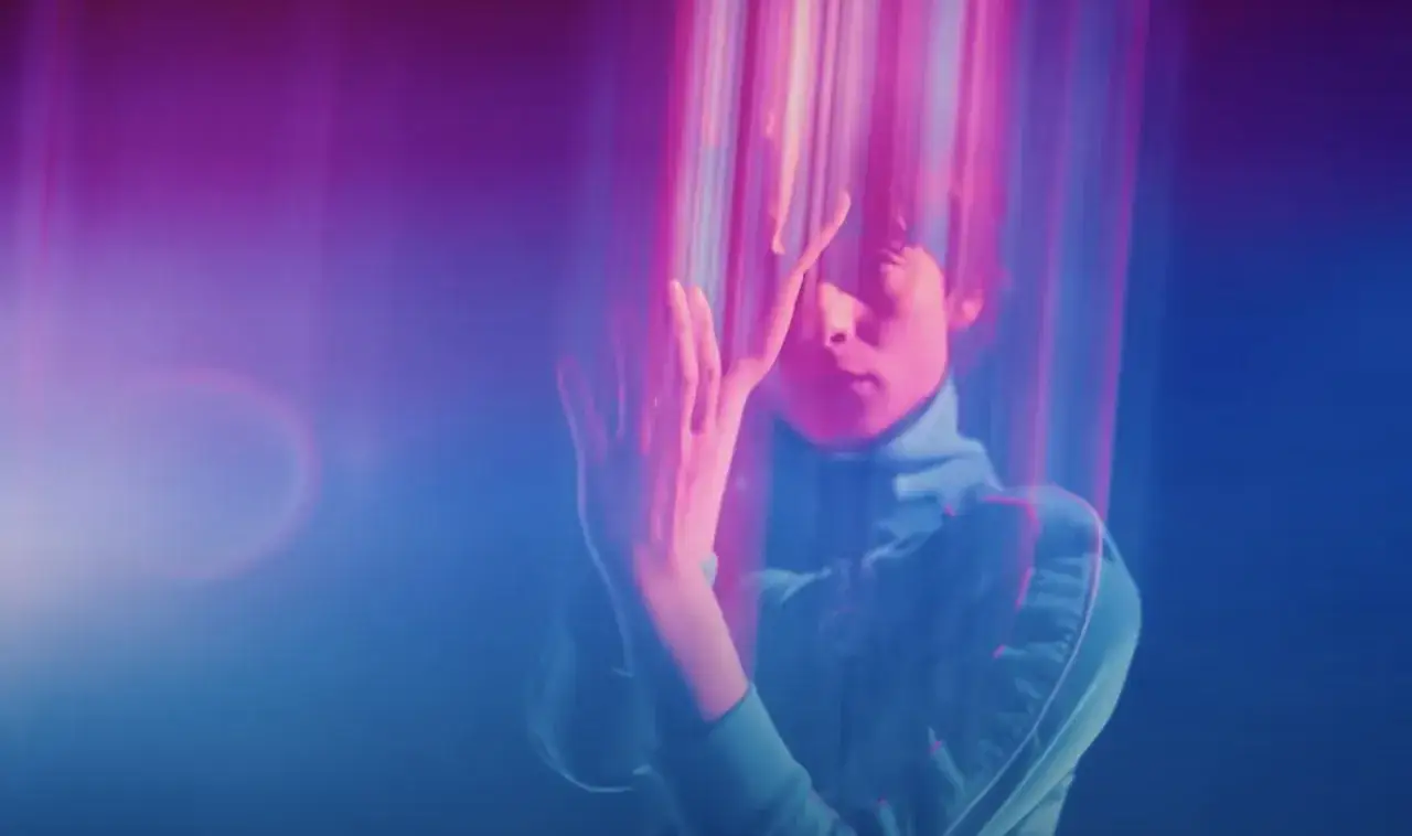 Un giovane con un dolcevita blu, immerso in luci rosa e blu vibranti, sembra un copenhagen cowboy in un sogno futuristico.
