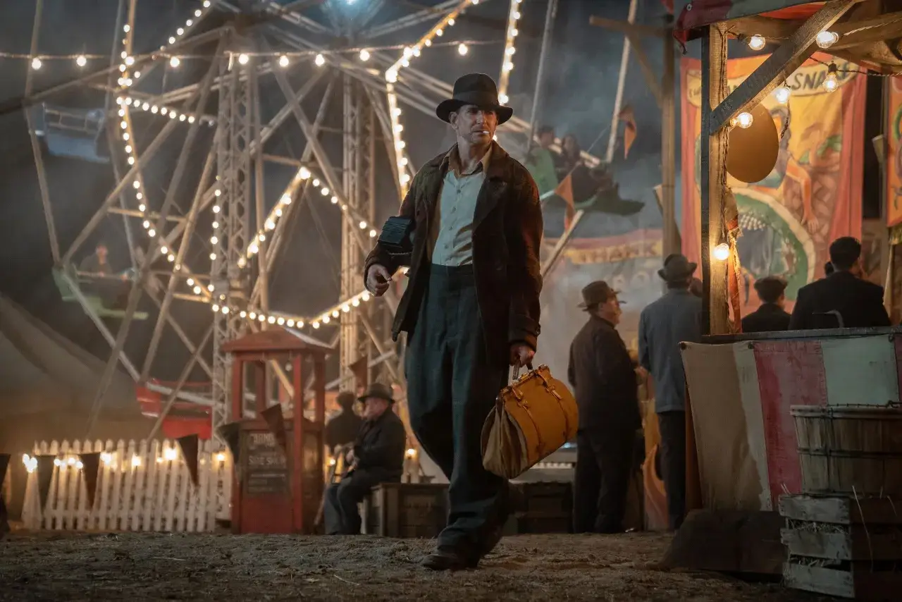Un uomo con un cappello cammina in una fiera delle illusioni illuminata da luci. Dietro di lui, una ruota panoramica e bancarelle creano un'atmosfera magica.