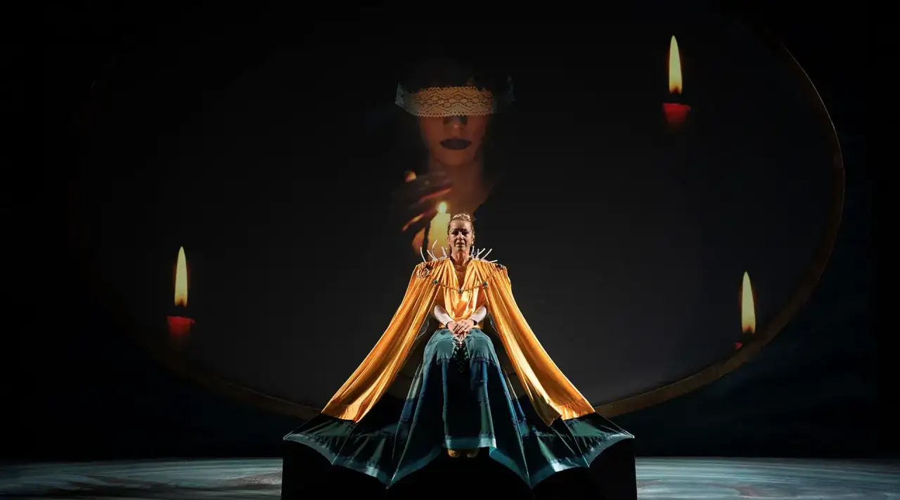 Scena teatrale con una donna in abito dorato e blu, circondata da candele. Sullo sfondo, un'immagine di una donna bendata evoca l'atmosfera del circe film.