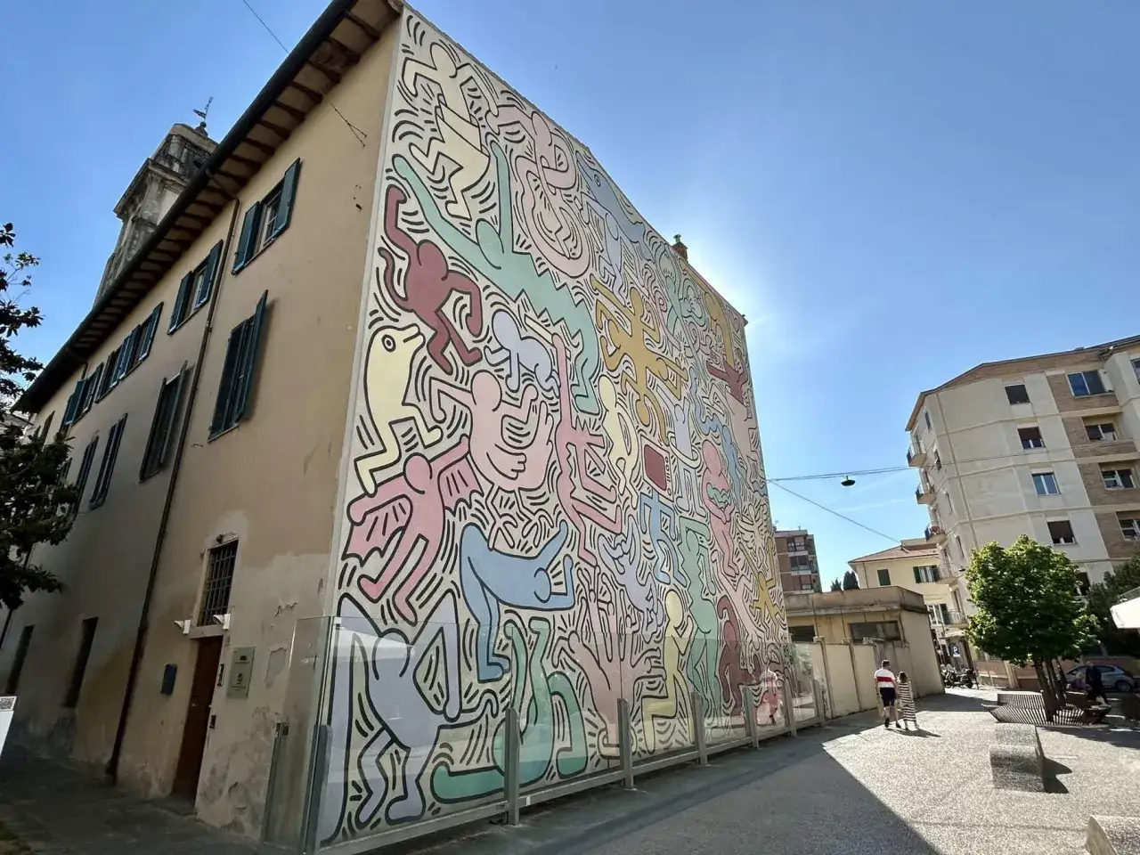 Un'enorme opera murale di Keith Haring, con le sue iconiche figure stilizzate in colori pastello, decora un edificio in una giornata di sole.