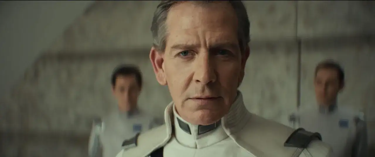 Ben Mendelsohn in uniforme, un volto noto nei film e programmi TV, con altri due uomini sfocati sullo sfondo.