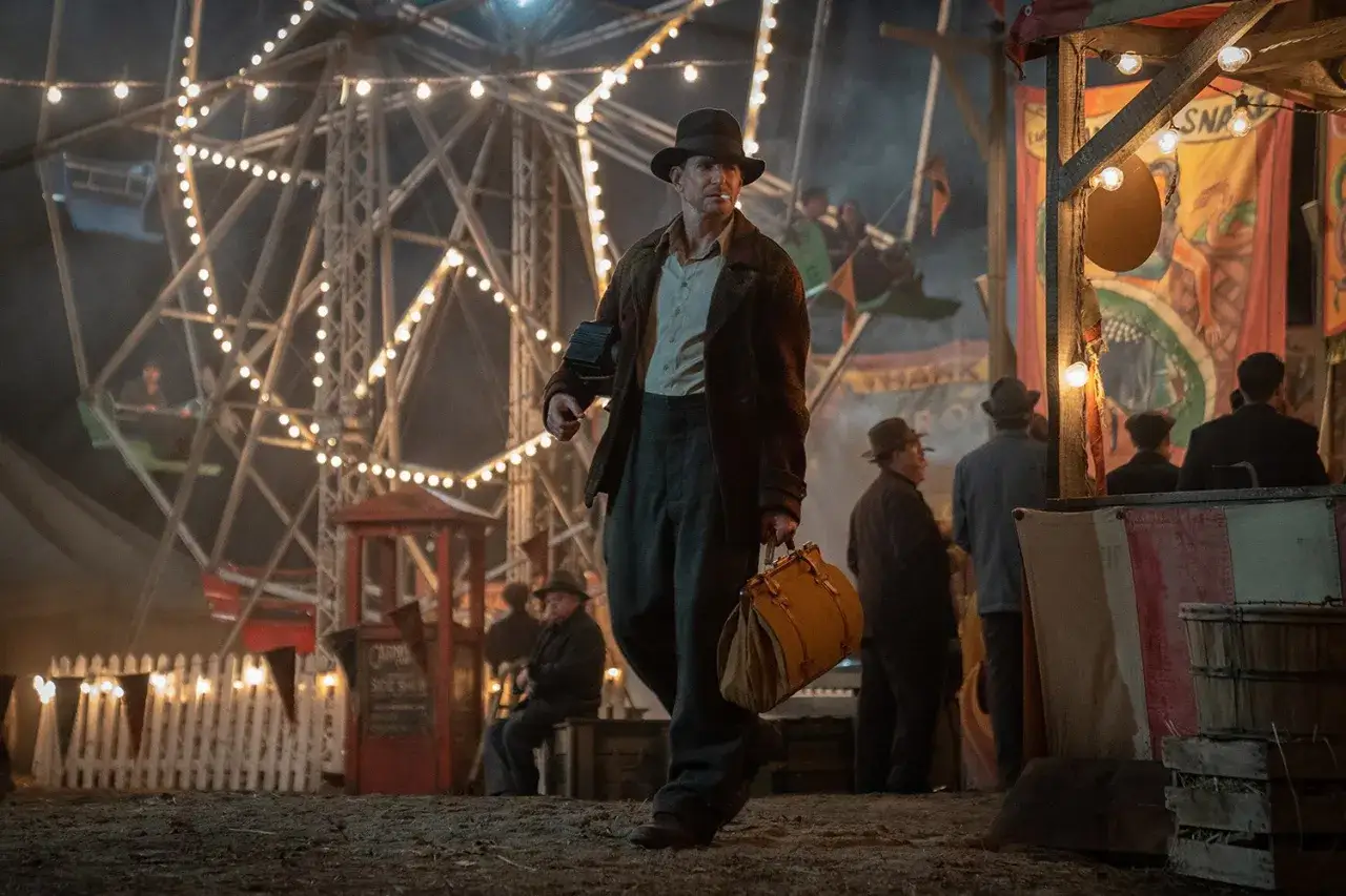 Un uomo con un cappello cammina in una fiera notturna illuminata da luci. La ruota panoramica sullo sfondo evoca