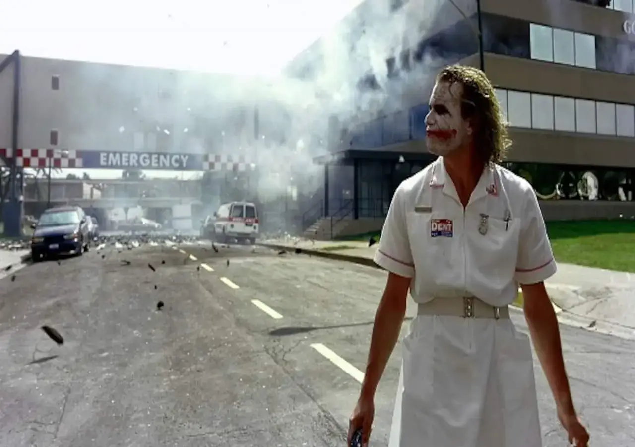 Il Joker, vestito da infermiere, osserva l'ospedale in fiamme dopo un'esplosione. Fumo e detriti ovunque.