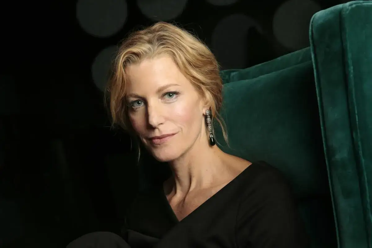 Skyler White di Breaking Bad, con capelli biondi e un orecchino pendente, seduta su una poltrona verde.