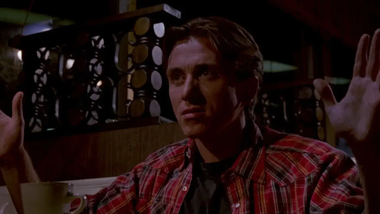 Mr. Orange, con camicia a quadri rossa, alza le mani in un gesto di resa o sorpresa, mentre una tazza è visibile in primo piano.