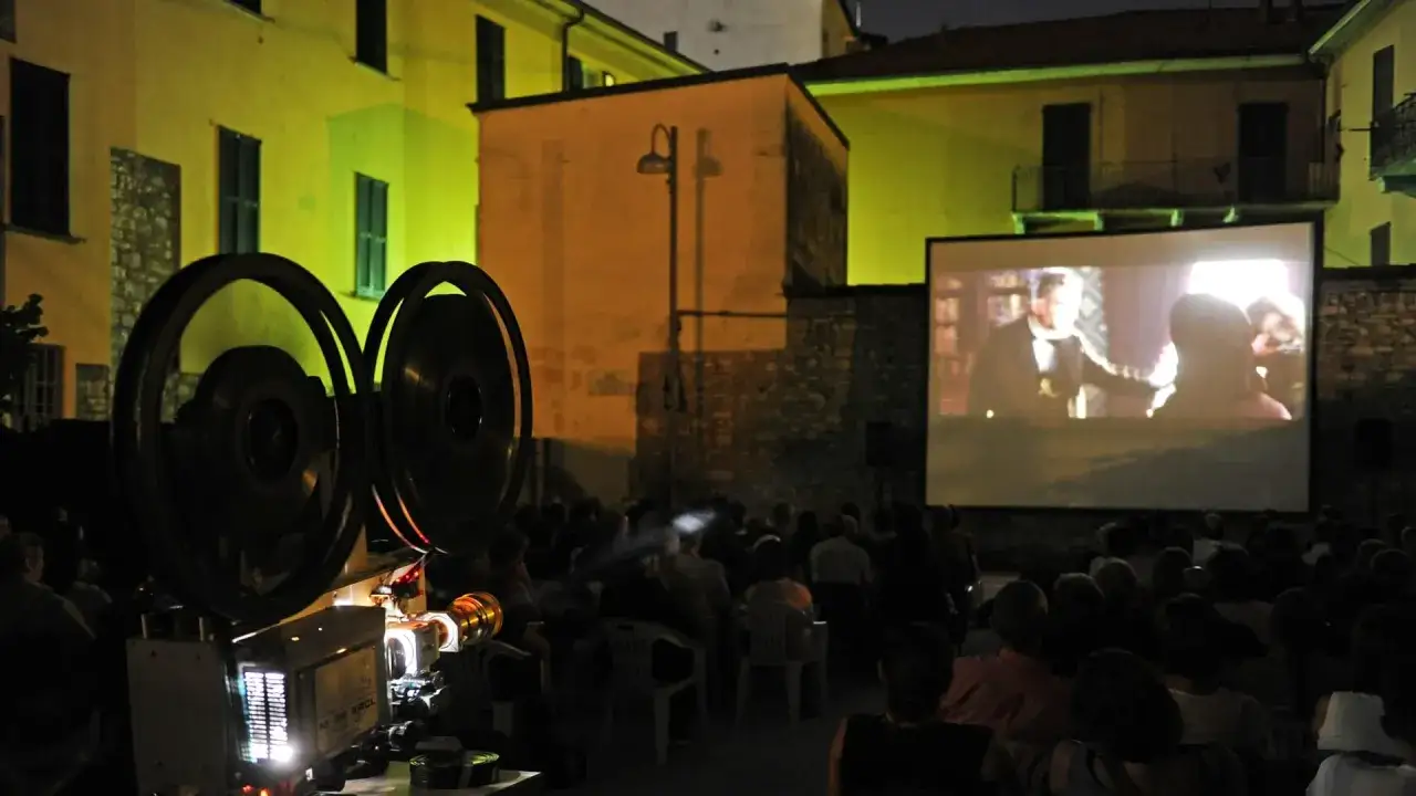 Un cinefilo felice assiste a una proiezione all'aperto, con un proiettore d'epoca e un pubblico seduto.