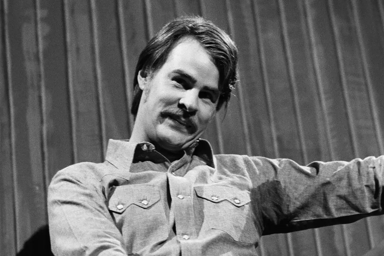 Dan Aykroyd giovane con baffi, indossa una camicia di jeans. Sorride con un'espressione furba.