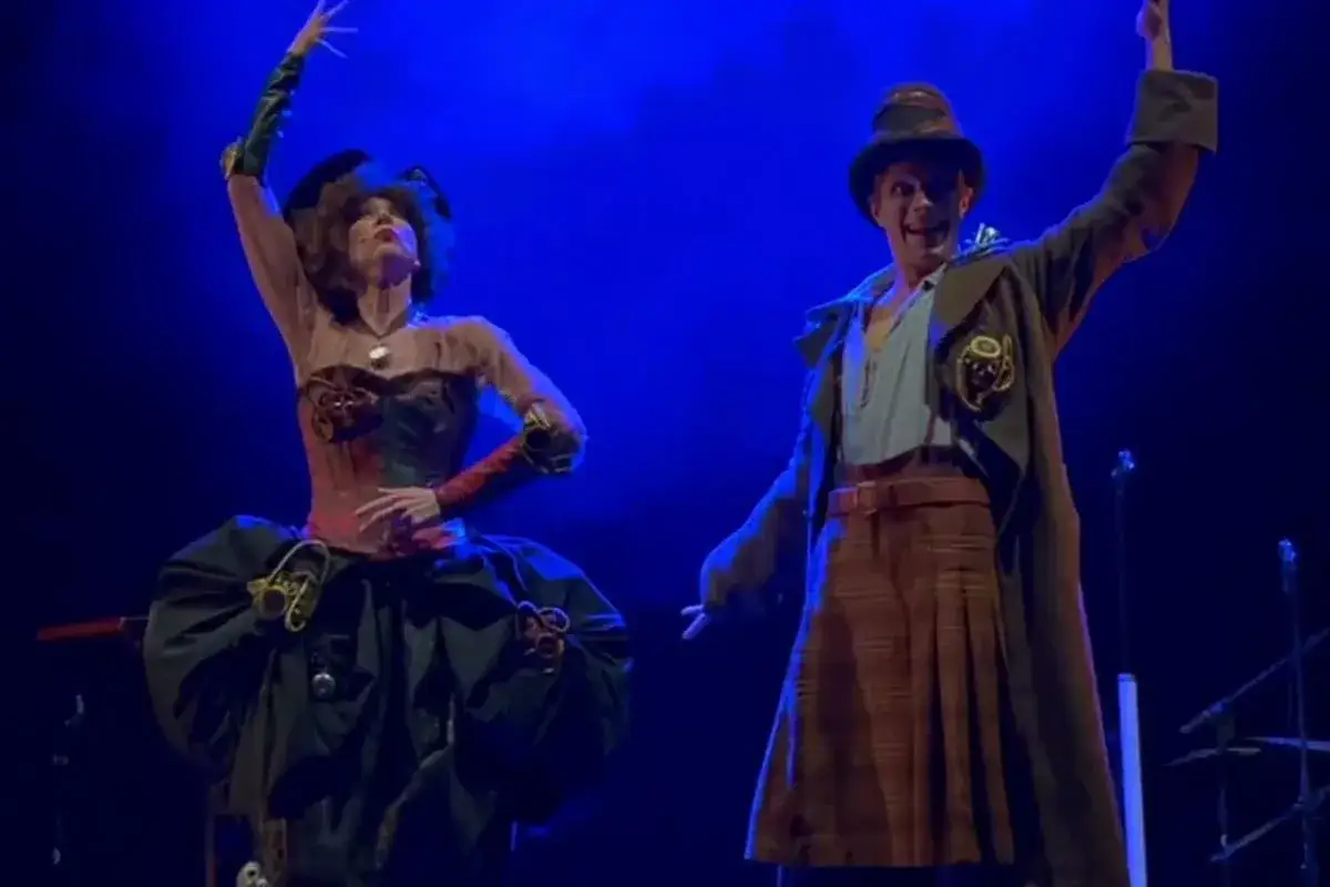 Due artisti in costume steampunk, uno con un abito elaborato e l'altra con un cappello e gonna a quadri, sul palco. Spiegazione de la doppia vita di Veronica.